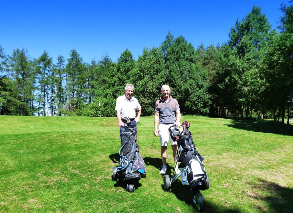 Two golfers standing on the course facing the viewer with their trolleys in front of them. They are smiling.