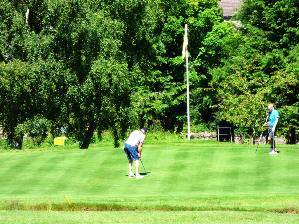 The green of a golf course. there are two players on the green. One is playing, the other stands to the side holding the flag.