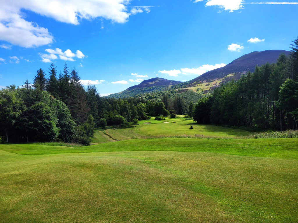 Looking down the fairway of a golf course. There are trees on either side of the grass and two hills overlooking.